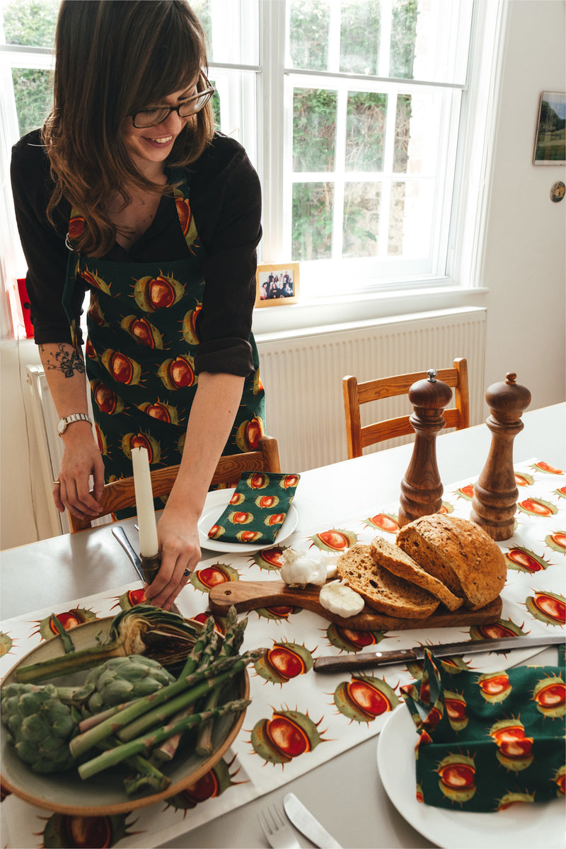 Lady smiling while setting the table with conker napkins and table runner. She is wearing conker apron. 