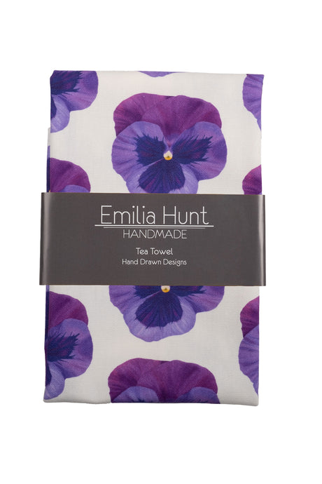 Folded tea towel with purple pansy design with card band across centre showing Emilia Hunt Handmade logo