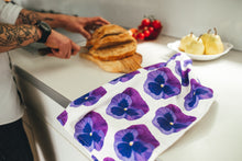 Load image into Gallery viewer, Purple pansy tea towel lying on kitchen worktop with man slicing bread in background
